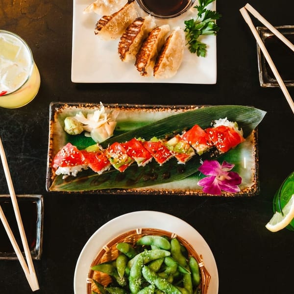 Japanese meal with sushi, gyoza, edamame, and two vibrant cocktails on a dark table.