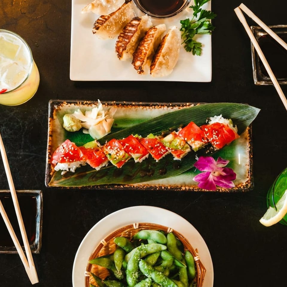 Sushi, gyoza, edamame, and two colorful cocktails arranged on a dark table with chopsticks.