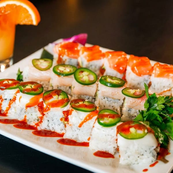 Assorted sushi rolls with salmon and jalapeños, garnished with parsley and a blue orchid, beside an orange cocktail.