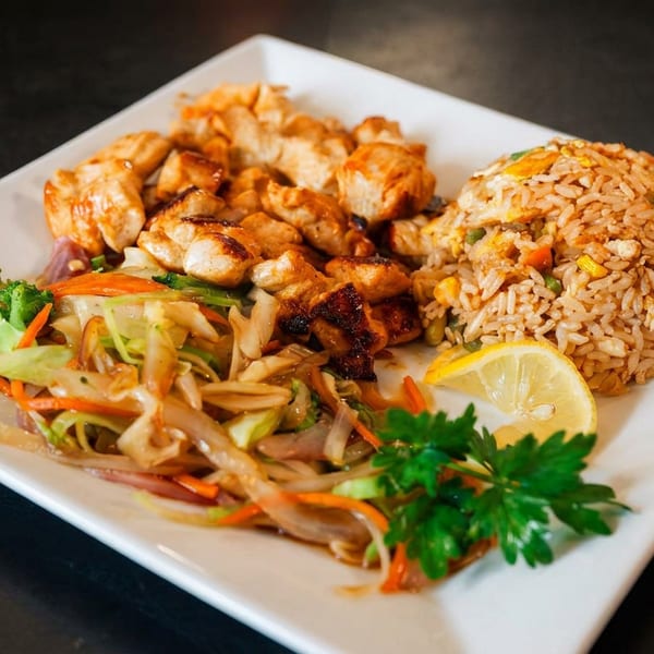 Teriyaki chicken, fried rice, and sautéed vegetables with a lemon slice on a white plate.