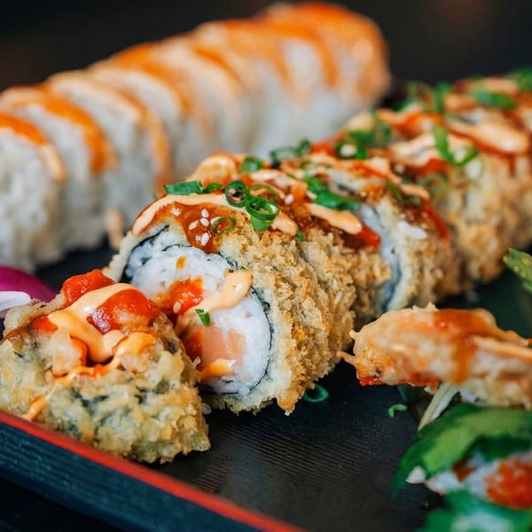 Tempura-fried sushi rolls with sauces, green onions, and sesame seeds, alongside another roll and a purple orchid.