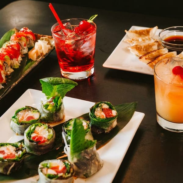 A vibrant spread of sushi, spring rolls, dumplings, and two cocktails on a dark restaurant table.